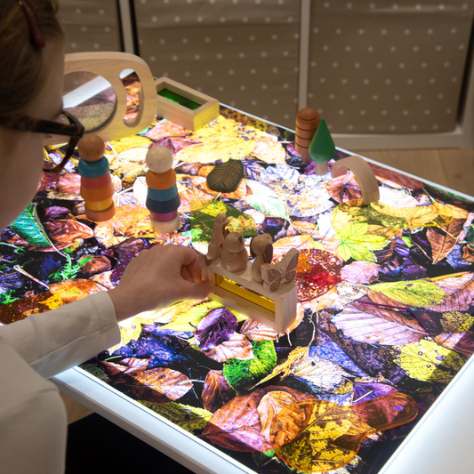 Person interacting with a colorful sensory table filled with leaf-shaped toys and a magnifying glass.