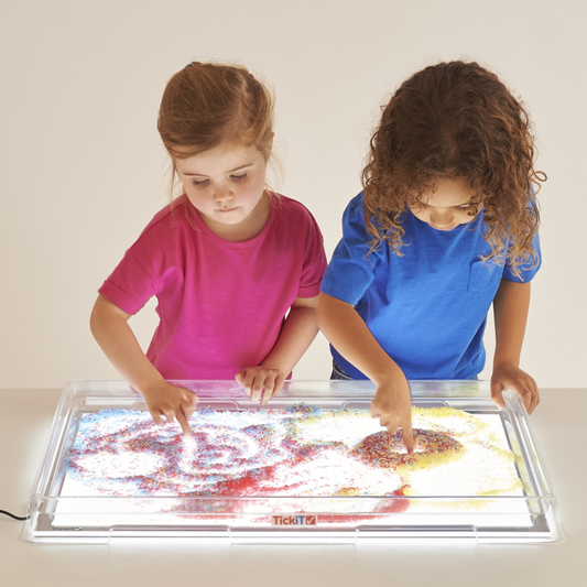 Two children playing with sensory rice on a tray over a light panel on a table.