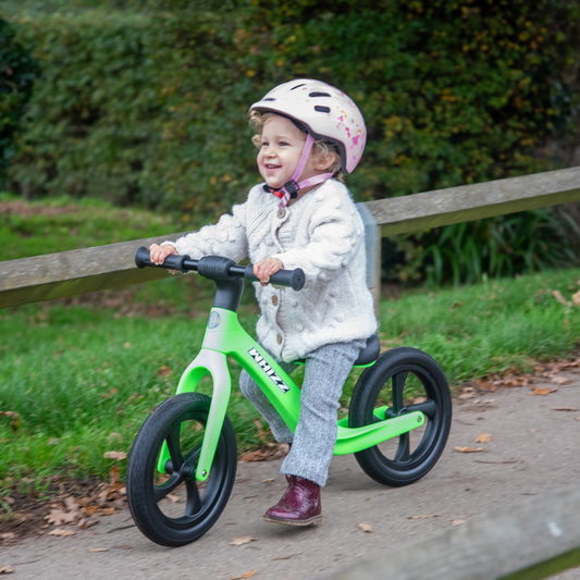 Child riding a green balance bike outdoors on a path with grass and trees in the background.
