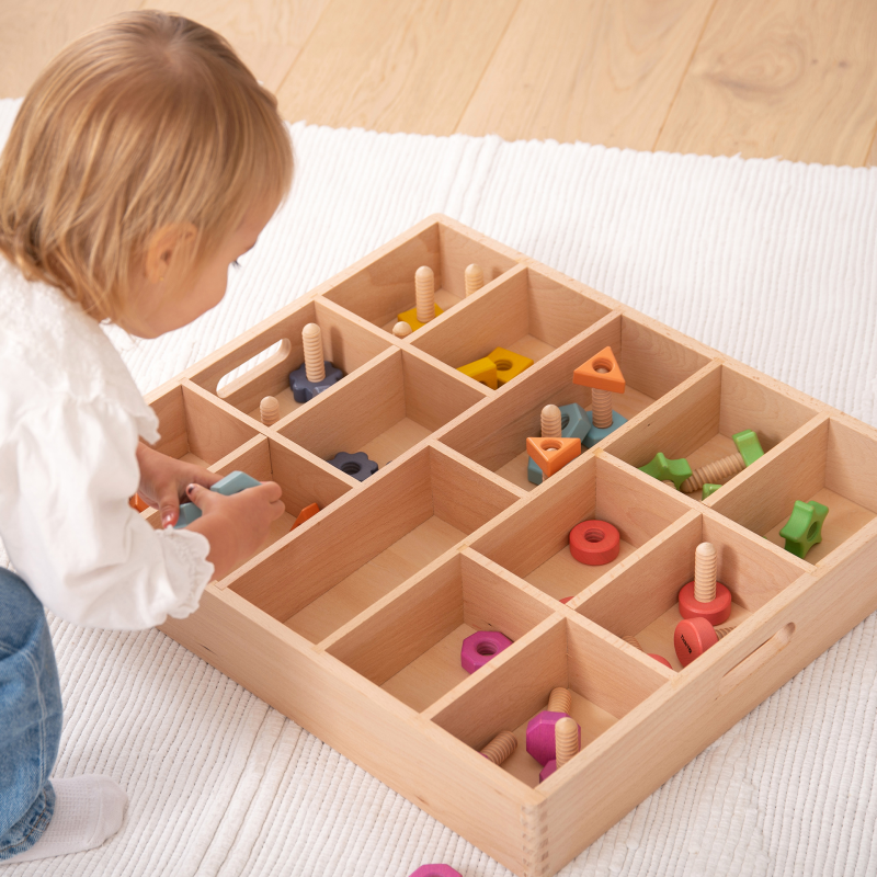 Child playing with wooden toys in a wooden sorting tray on a light-coloured floor.
