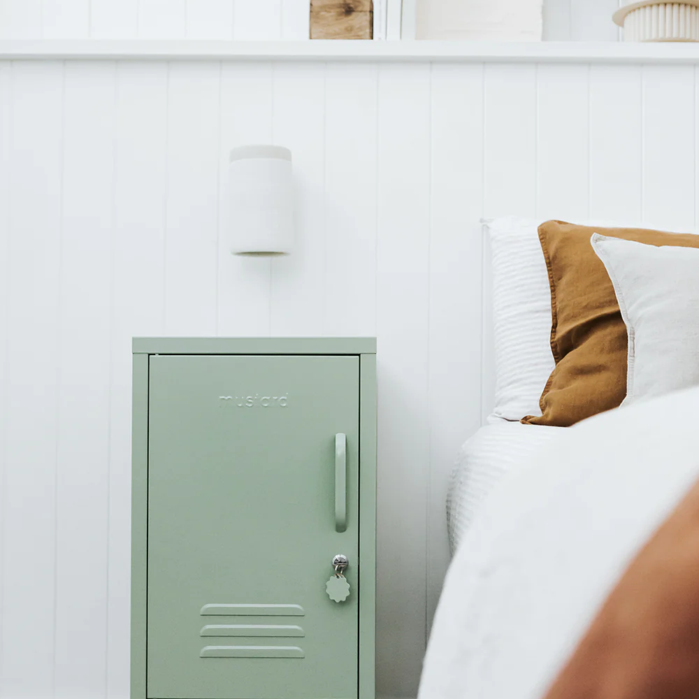 Green metal locker against a white wall with a bed and decorative items in the background