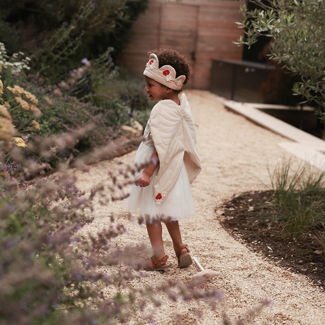 Child in a white dress and floral headband standing on a gravel path with greenery around.