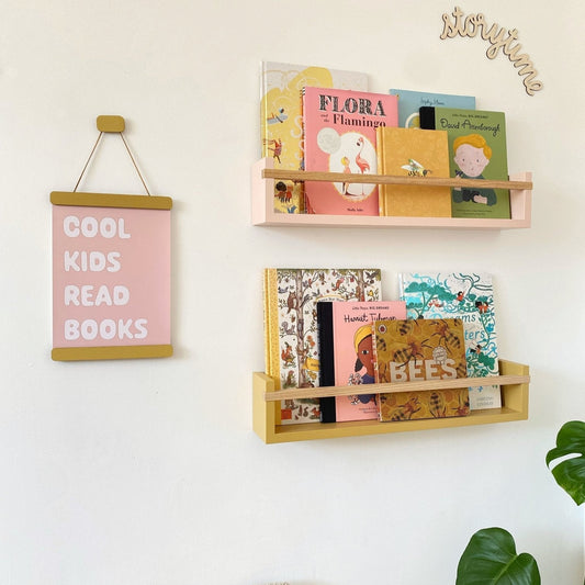 Wooden bookshelf with children's books on a white wall, next to a pink sign with text.