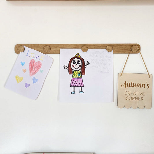 Children's drawings and a sign on a wooden peg rack against a white wall.