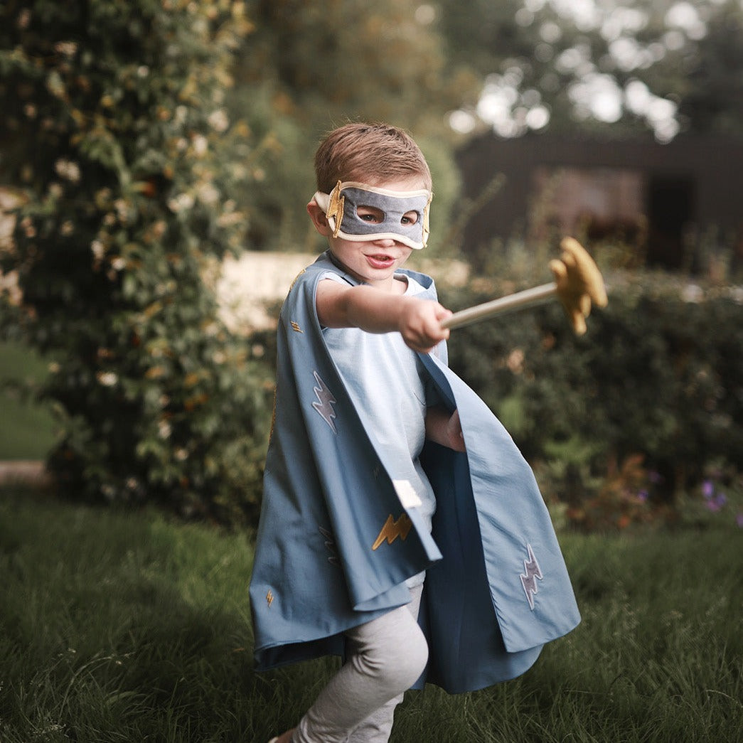 Child in superhero costume with cape and mask, holding a toy sword outdoors.