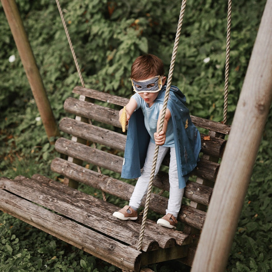Child in superhero costume on a swing set with greenery in the background